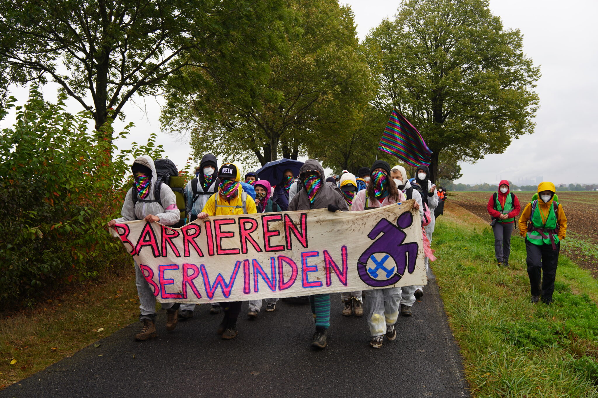 Mehrere Menschen laufen über einen Feldweg. Sie halten ein Transparent mit der Aufschrift "Barrieren Überwinden" und einem Rollstuhl-Pictogramm in der Hand.