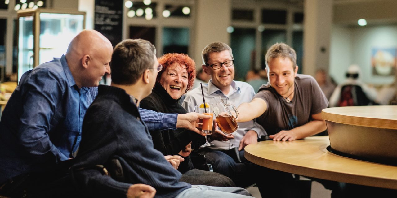 Eine Gruppe von Menschen mit Behinderungen sitzt an einem Tisch und stößt mit Getränken an.